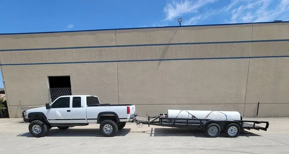 White pickup truck towing a trailer with a large white tank, parked next to a tan building.
