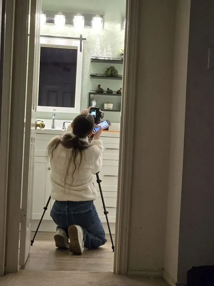 Person kneeling, taking photo of bathroom. Camera on tripod, doorway frame, white walls, vanity, mirror.