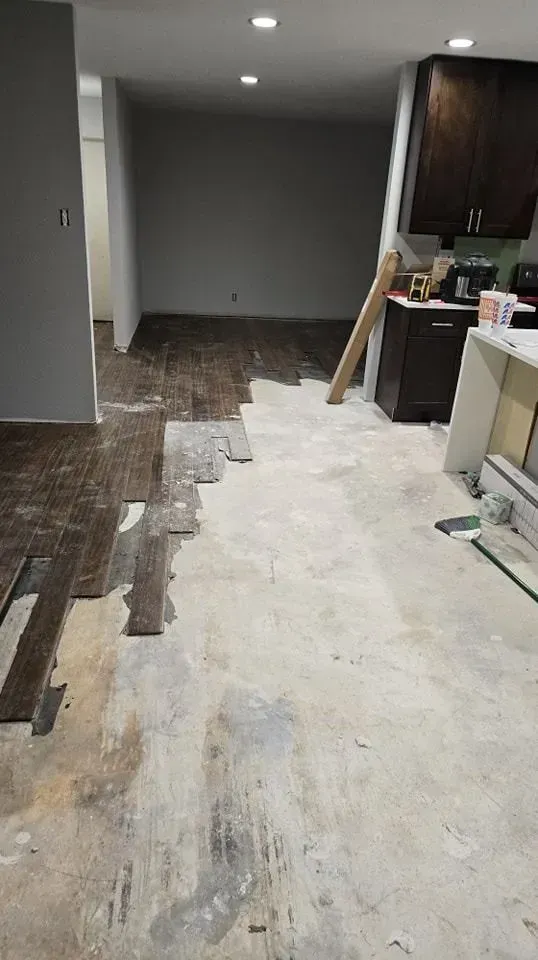 Interior view of a room under renovation. Old flooring partially removed, exposing concrete subfloor. Kitchen cabinets in the background.