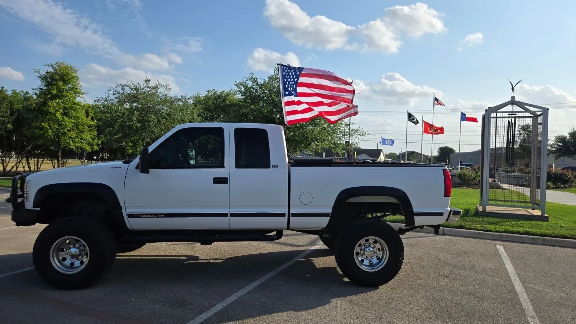 White lifted pickup truck with American flag in the back, parked outdoors.