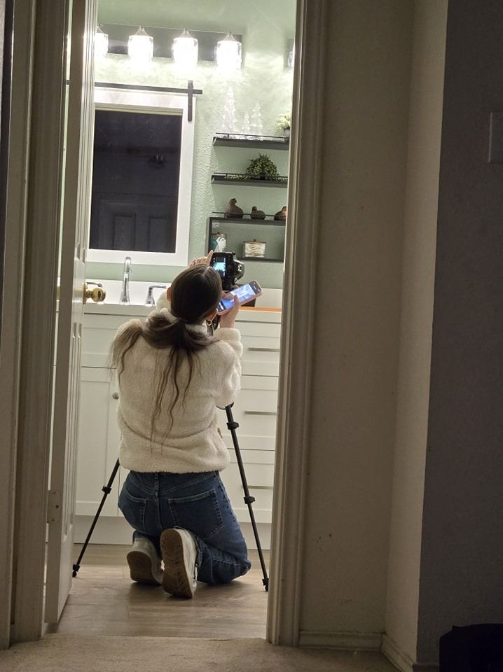 Person on a tripod, photographing a bathroom. They are kneeling in a doorway.