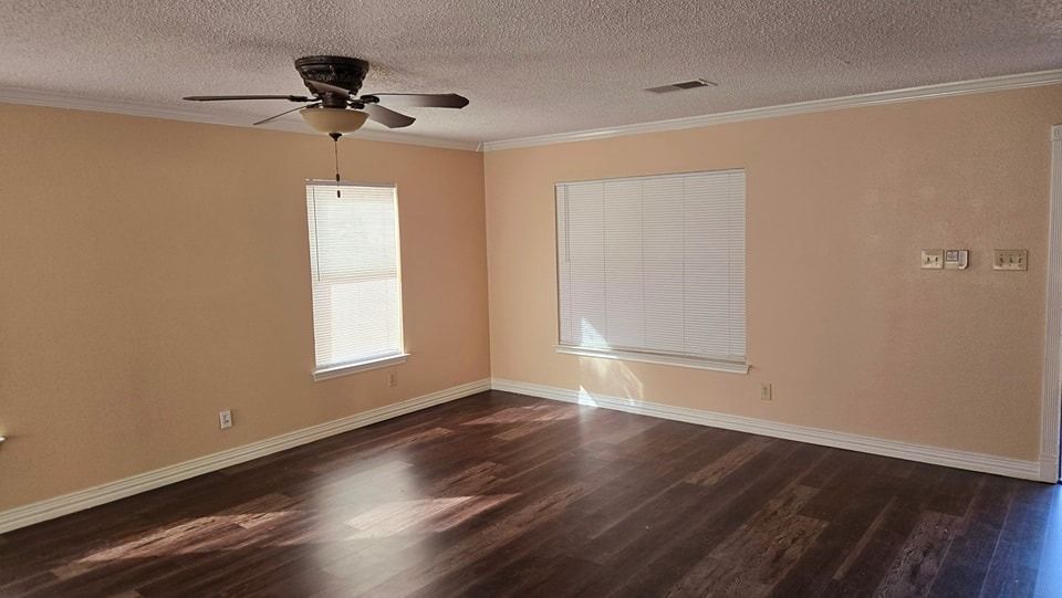 Empty living room with wood flooring, beige walls, two windows with blinds, and a ceiling fan.