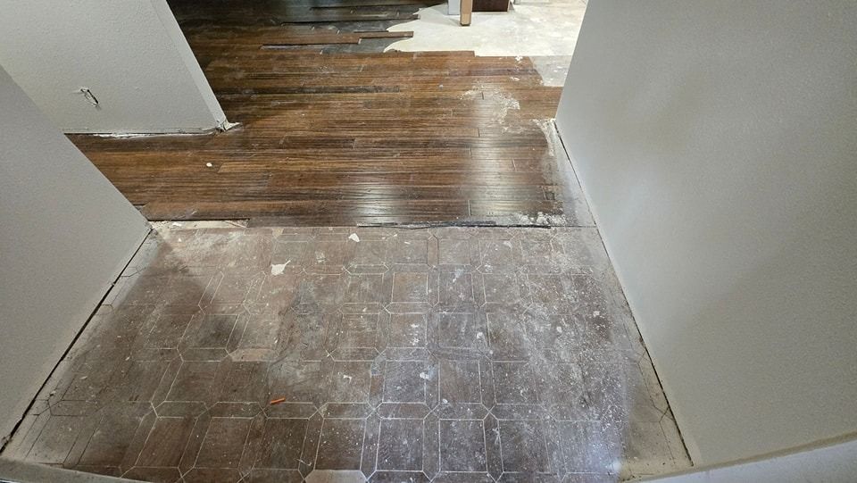 Hallway with damaged tile floor transitioning to dark hardwood flooring. Walls are white, construction in progress.