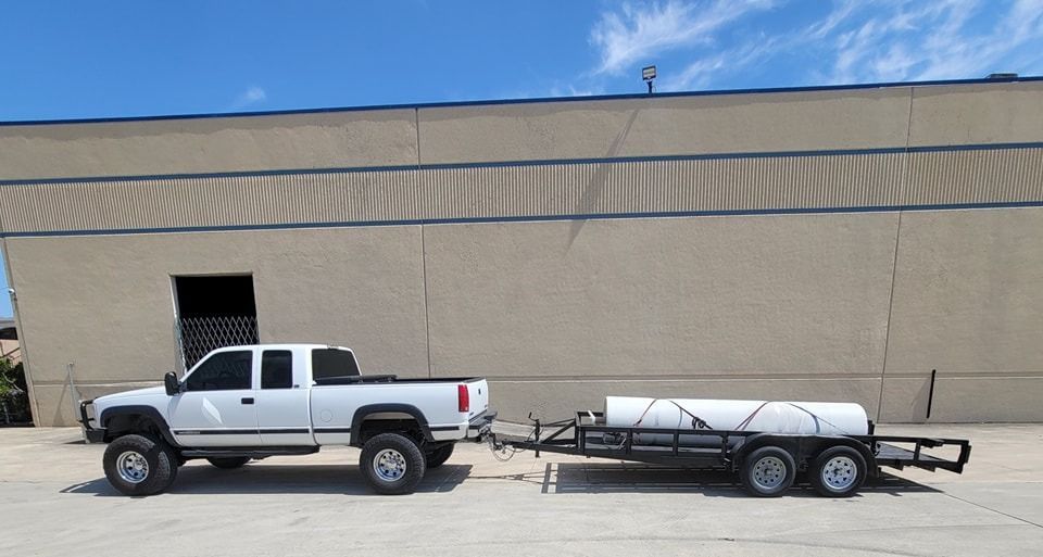 White pickup truck towing a trailer with a large white tank, parked in front of a building on a sunny day.