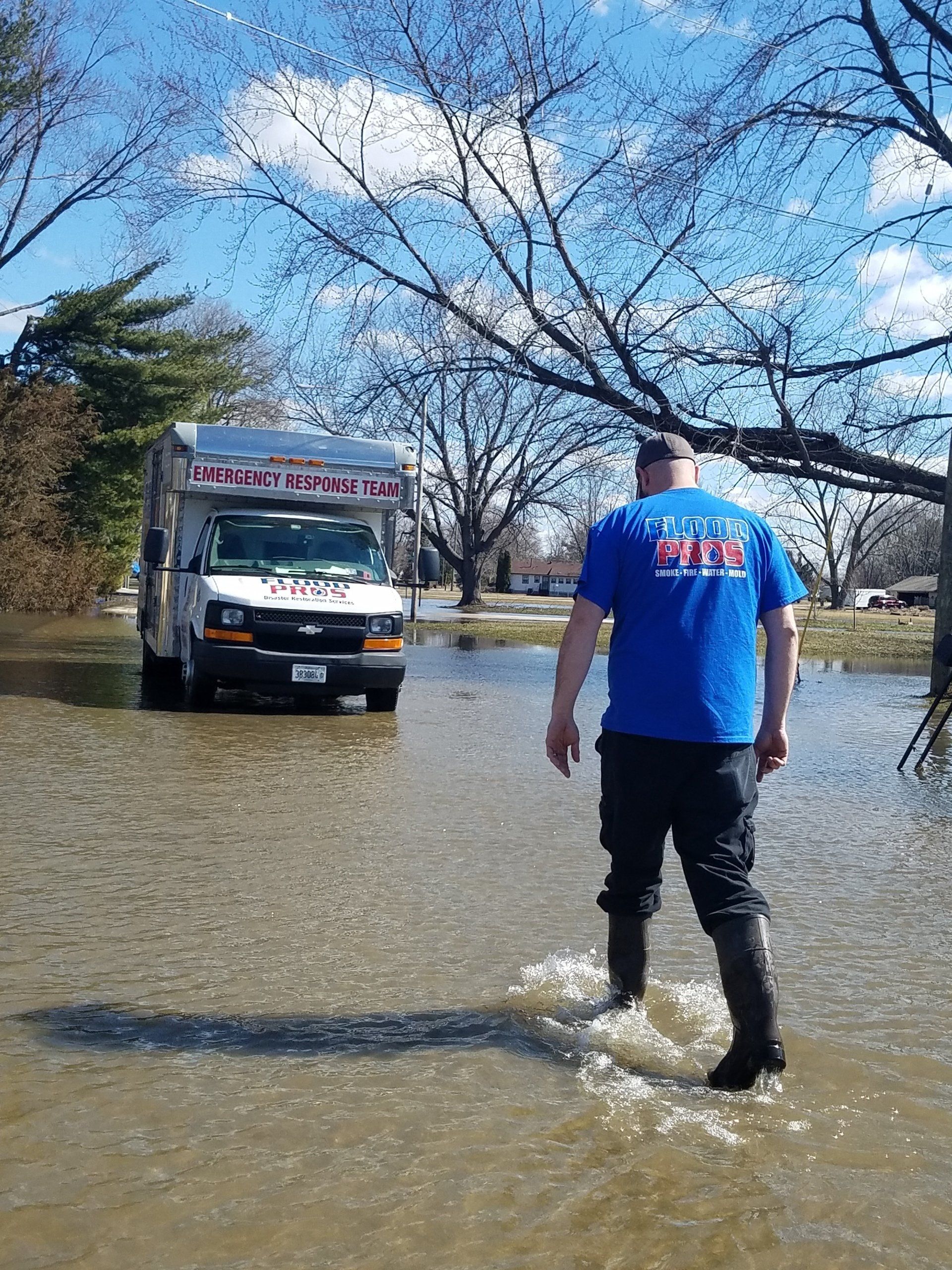 Man in waders walks toward a service van through floodwater. Blue sky with trees and houses in the background.