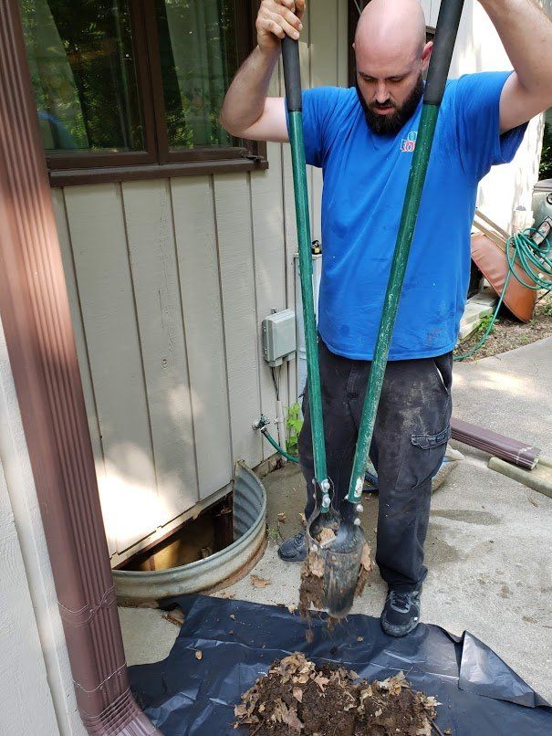 A man digs with a post hole digger near a building. He wears a blue shirt and is focused.