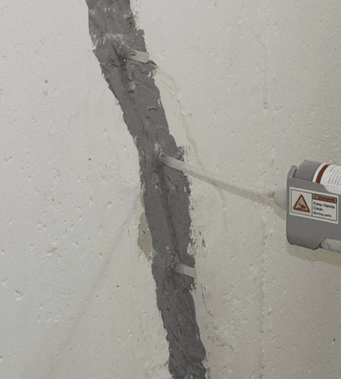 White wall with a vertical crack being repaired. A tube is injecting material into the crack, held in place by clips.