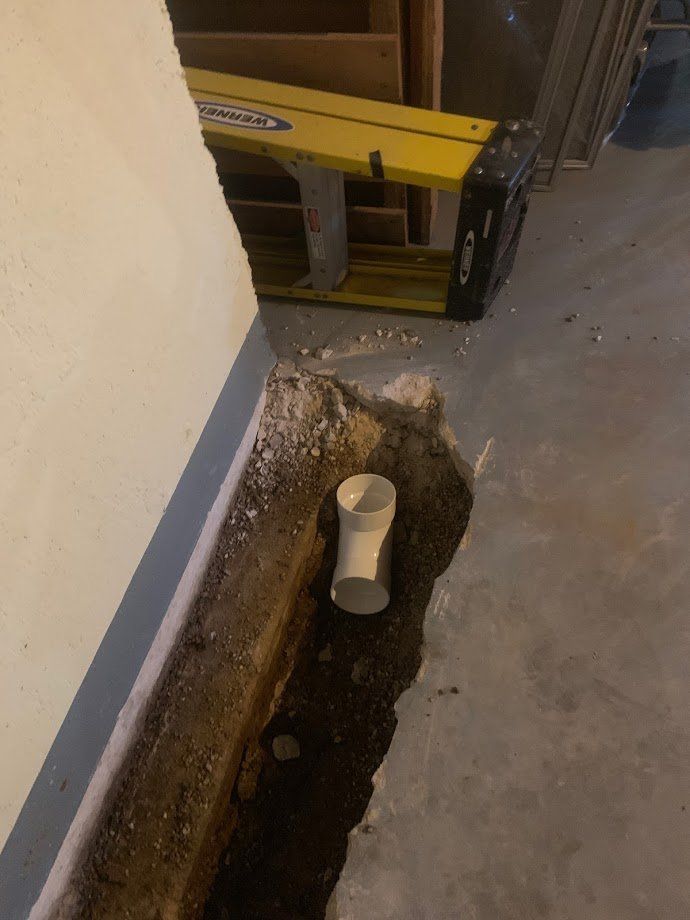 A trench dug in a concrete floor reveals a white PVC pipe. A yellow ladder rests nearby.