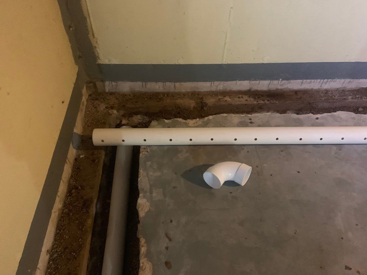 A white perforated pipe and a white elbow fitting are on a concrete floor near a wall, likely for drainage.