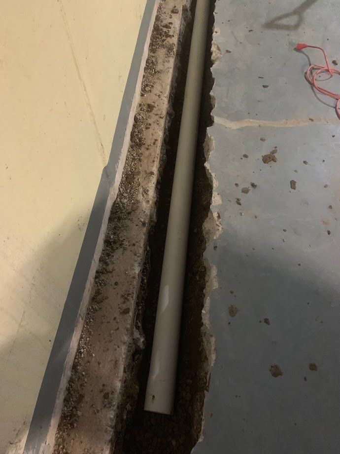 A gray pipe lies in a trench cut into a gray concrete floor, next to a light yellow wall.