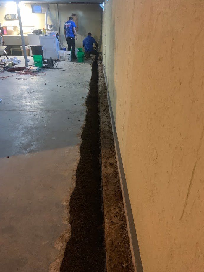 Workers digging a trench along a basement wall. A concrete floor, tools, and other equipment are visible in the background.