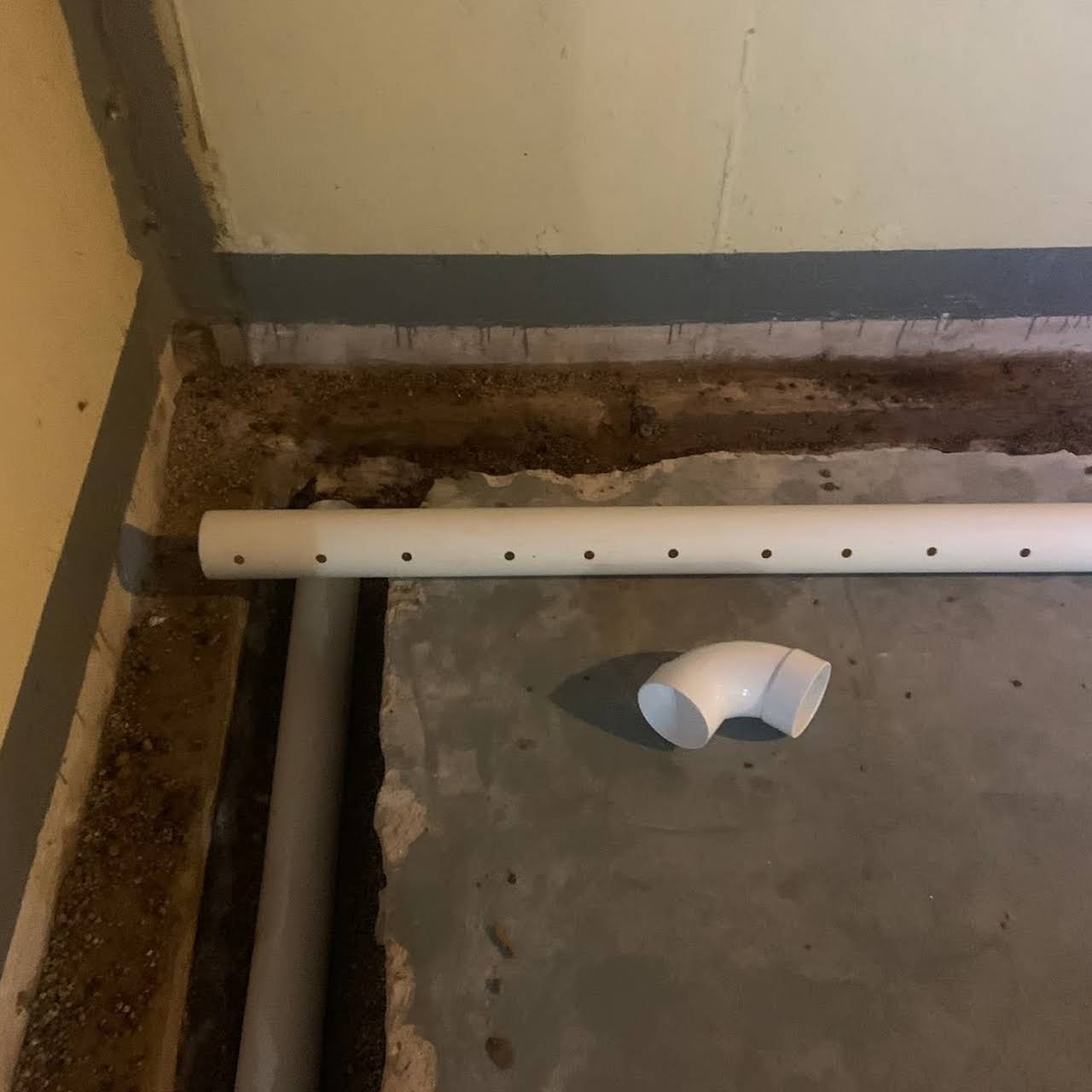 White perforated pipe and elbow on a concrete floor in a basement. A gray pipe runs alongside the white pipe near the foundation.