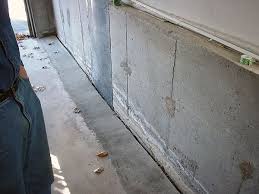 Cracked concrete basement wall with water damage.  A person stands nearby. The wall is gray with vertical and horizontal cracks.