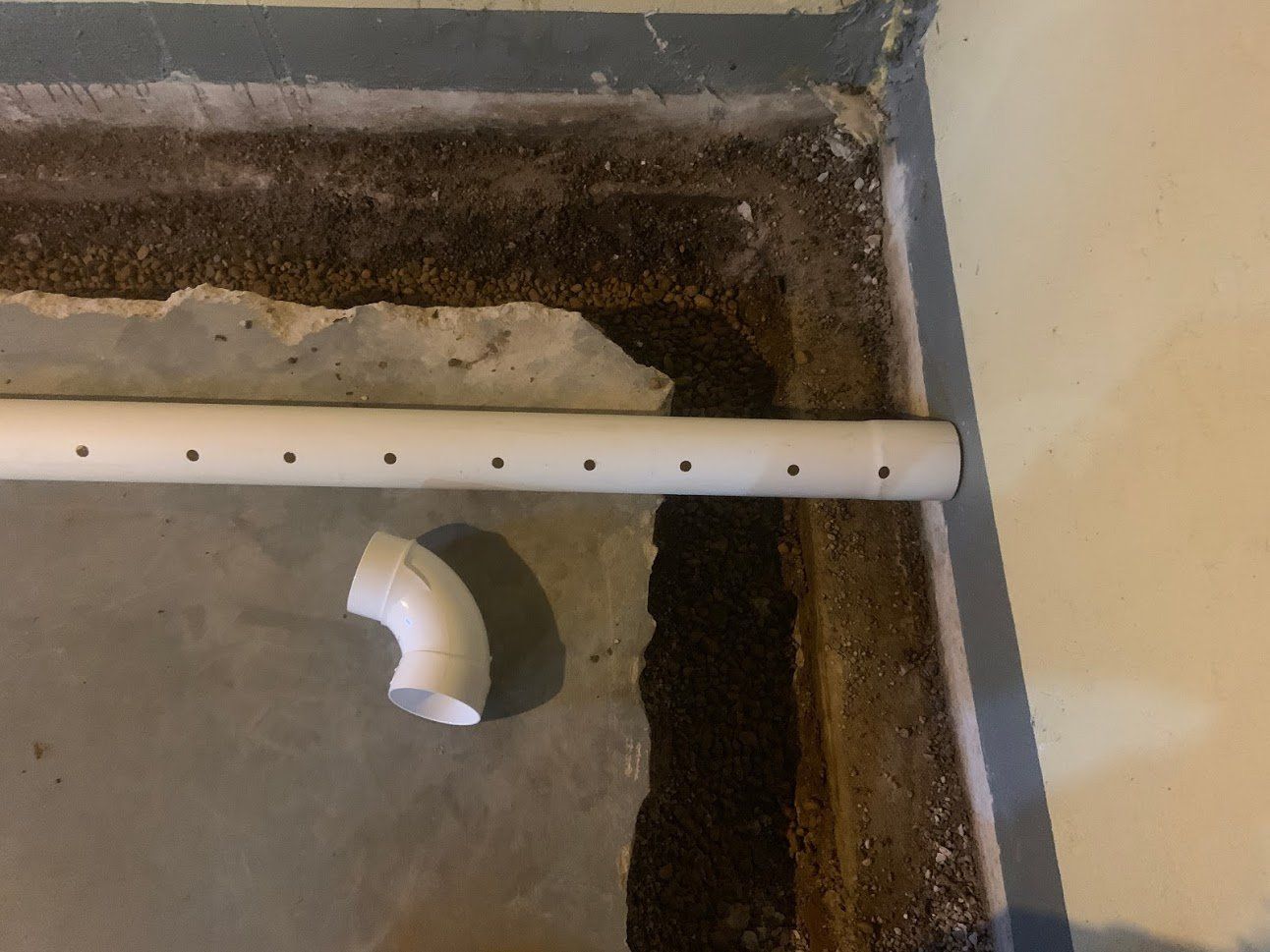 A white PVC pipe with evenly spaced holes lies on a concrete floor, next to an elbow joint and a trench.