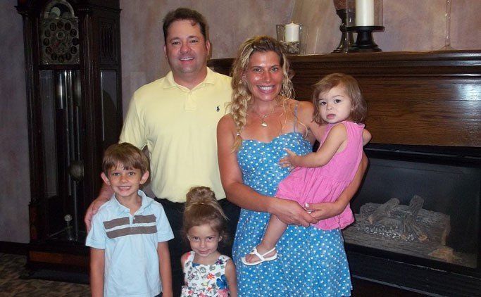 A family of five poses in front of a fireplace. Parents smile, holding their two young daughters while their son stands to the left.