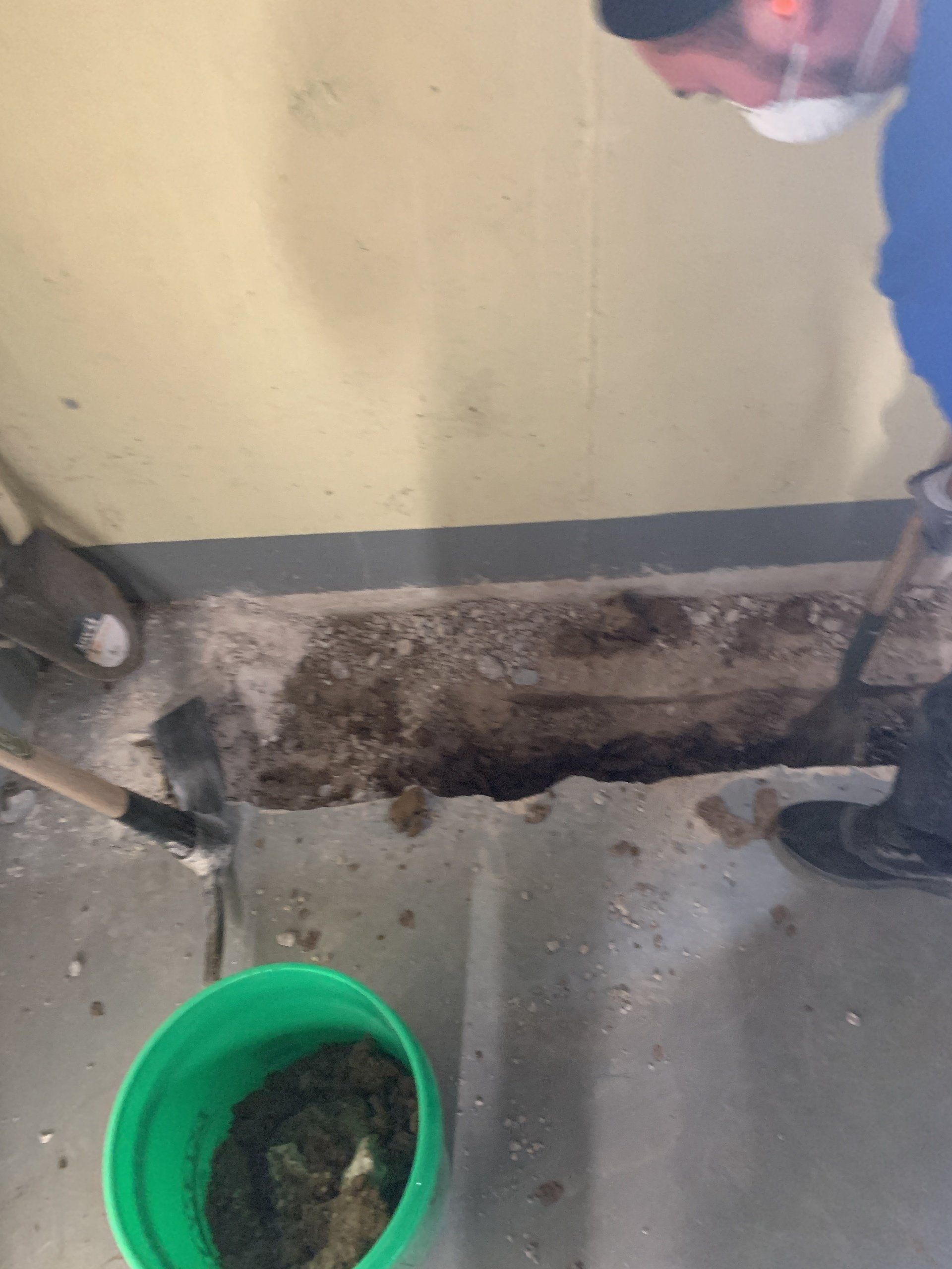 Person in a mask shoveling debris from a floor, green bucket with waste, and wall are visible.