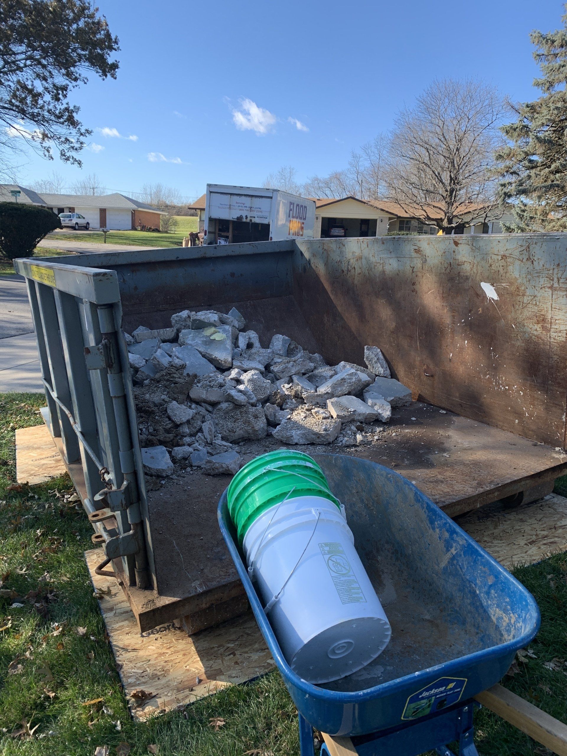 A wheelbarrow with two white buckets in front of a large dumpster filled with broken concrete, outdoors on a sunny day.