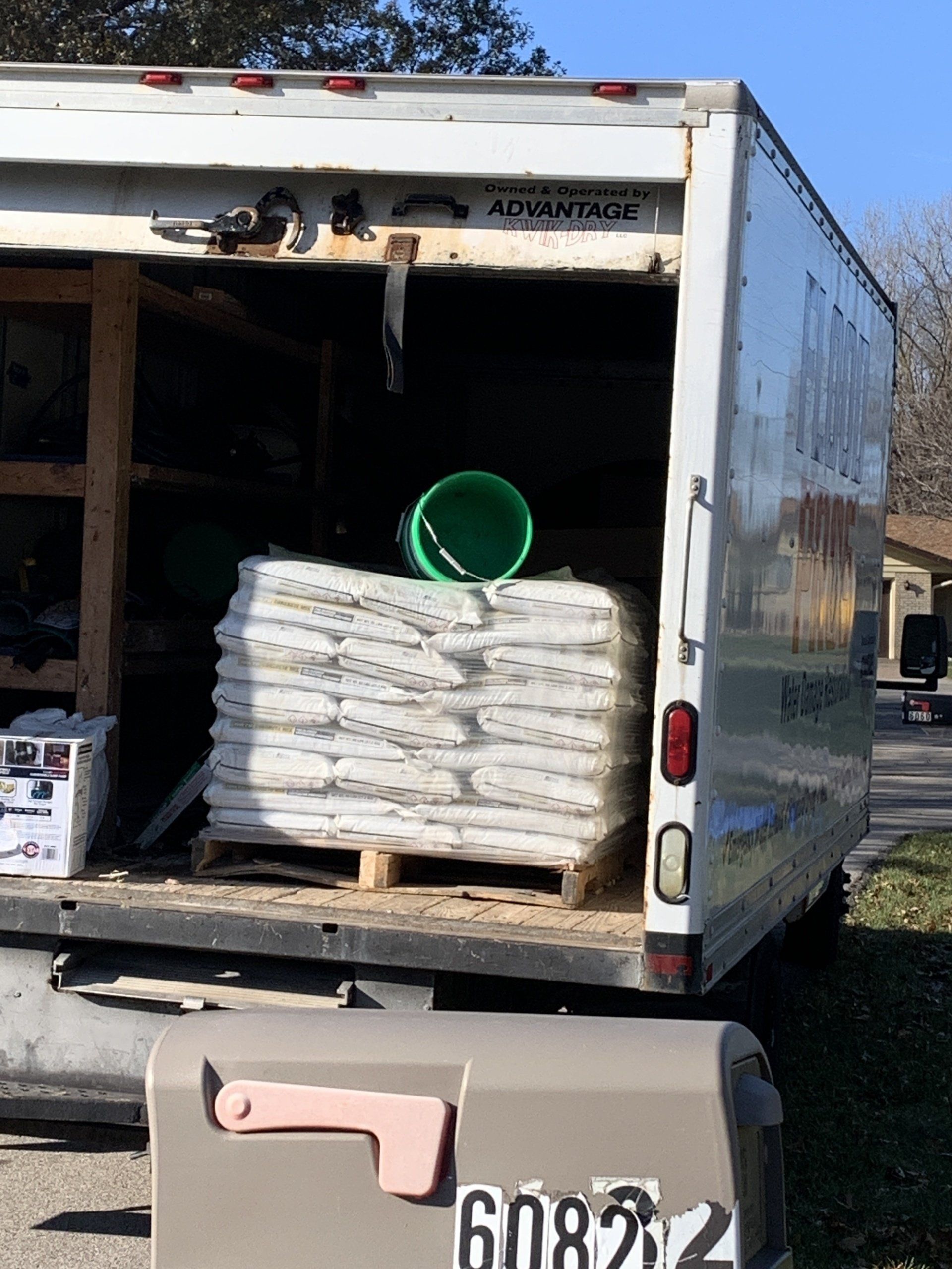 White truck bed loaded with stacked bags, green bucket, and a mailbox with house number 6082 in front.