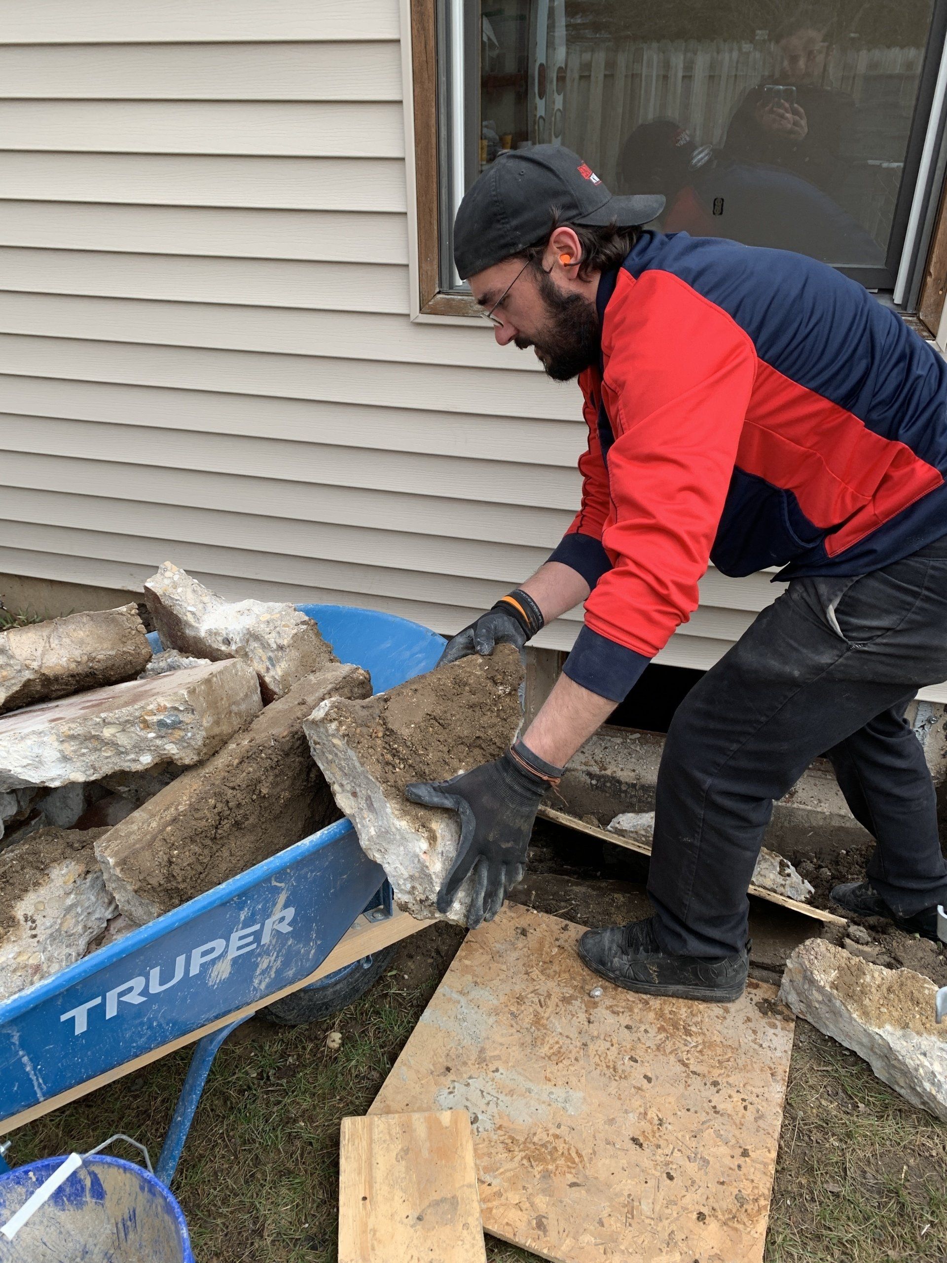 A man in a red and blue jacket and black gloves loads concrete debris into a blue wheelbarrow near a house.