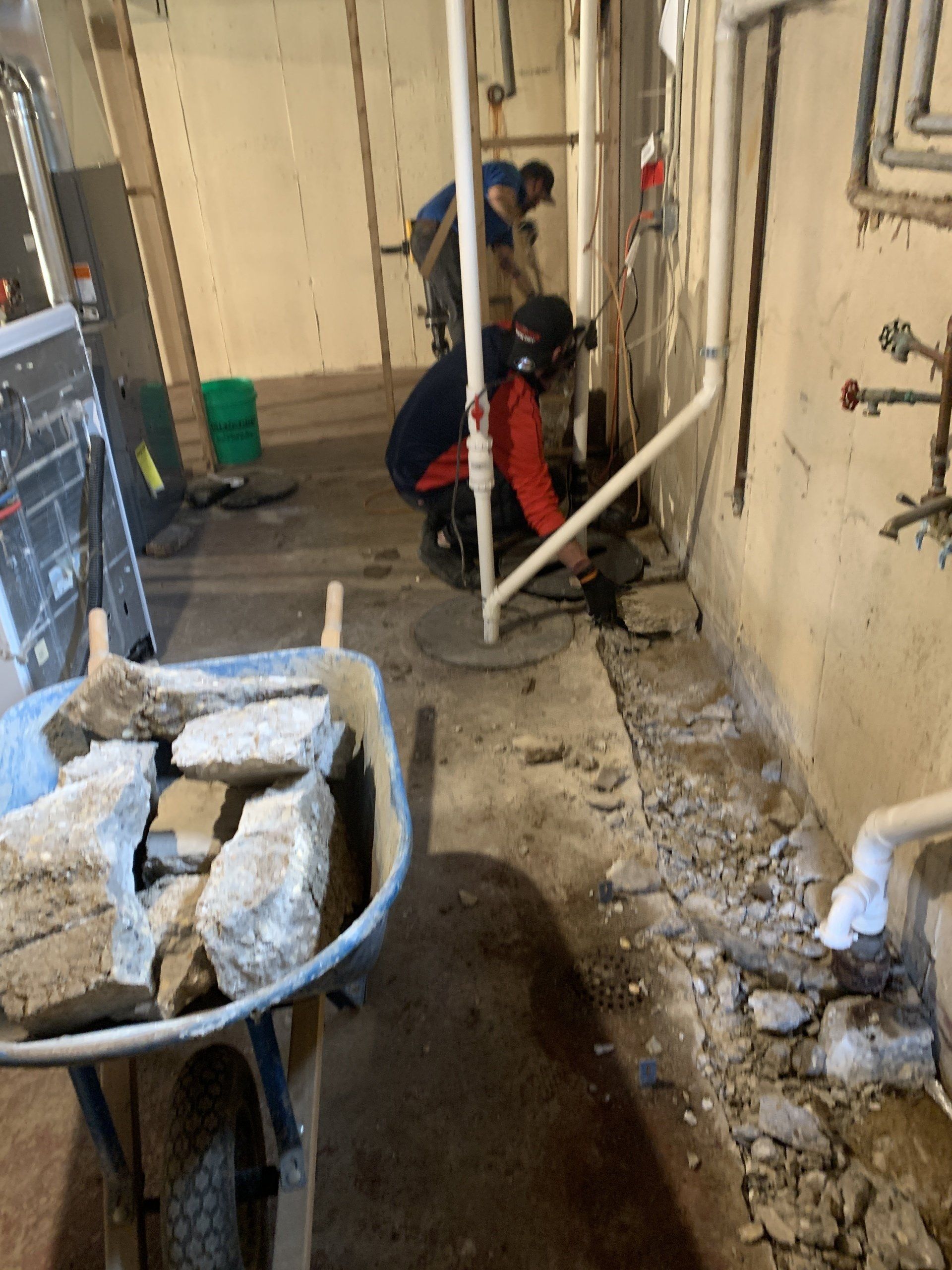 Three construction workers are cutting into a concrete floor next to a wall. A wheelbarrow of debris is in the foreground.