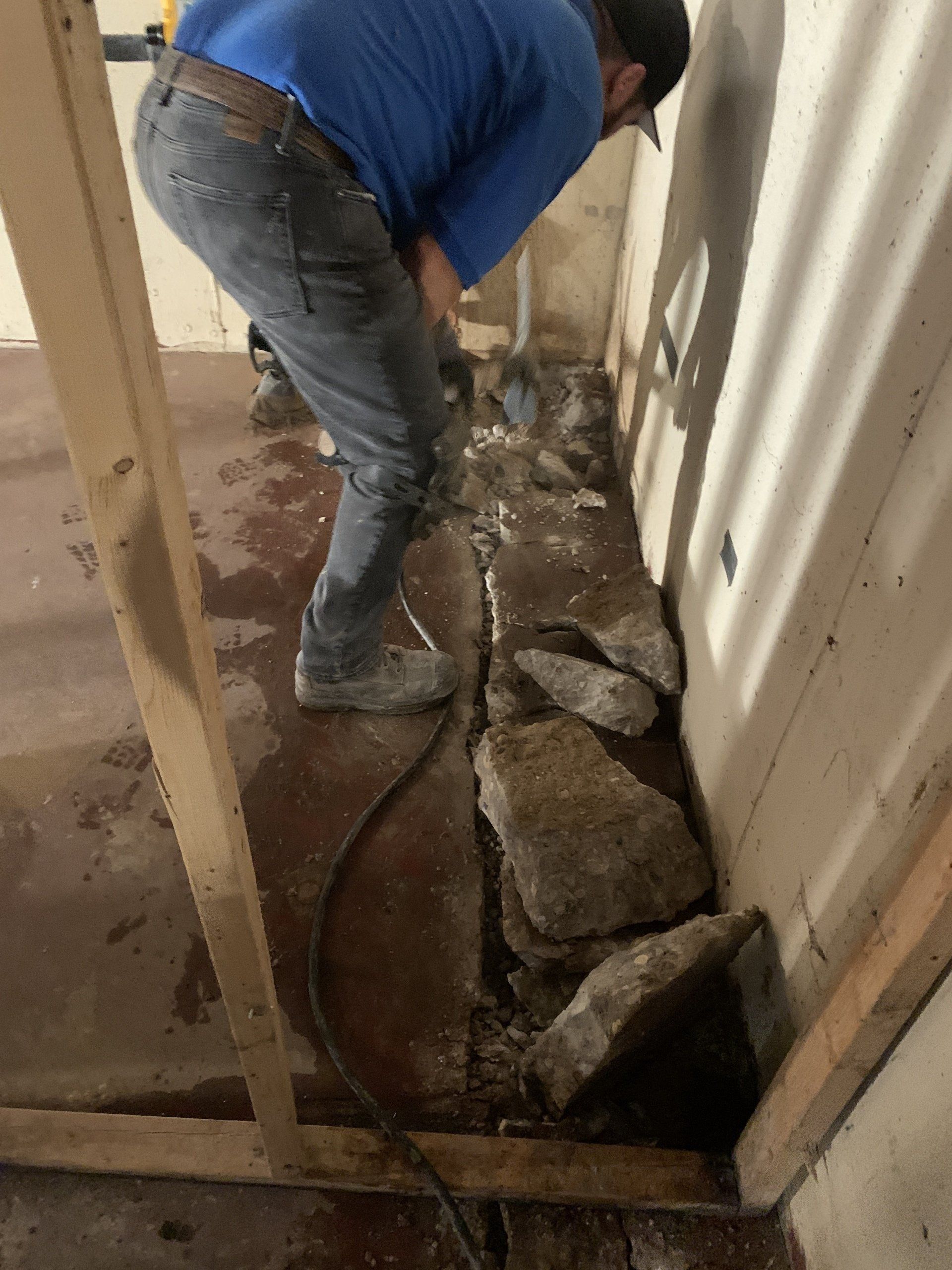 Person jackhammering concrete near a wall. They wear jeans, work boots, and a blue shirt, standing in a narrow room.