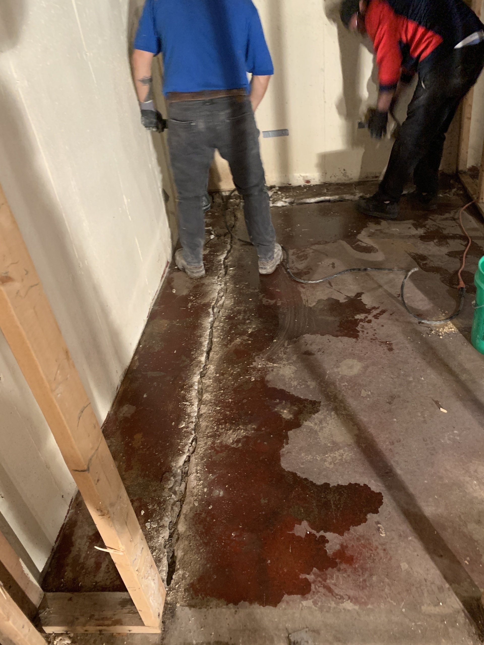 Two people in a basement working on a cracked concrete floor. Brown-stained concrete floor is wet, and debris scattered. One person holds a tool, another is using a tool on the floor.