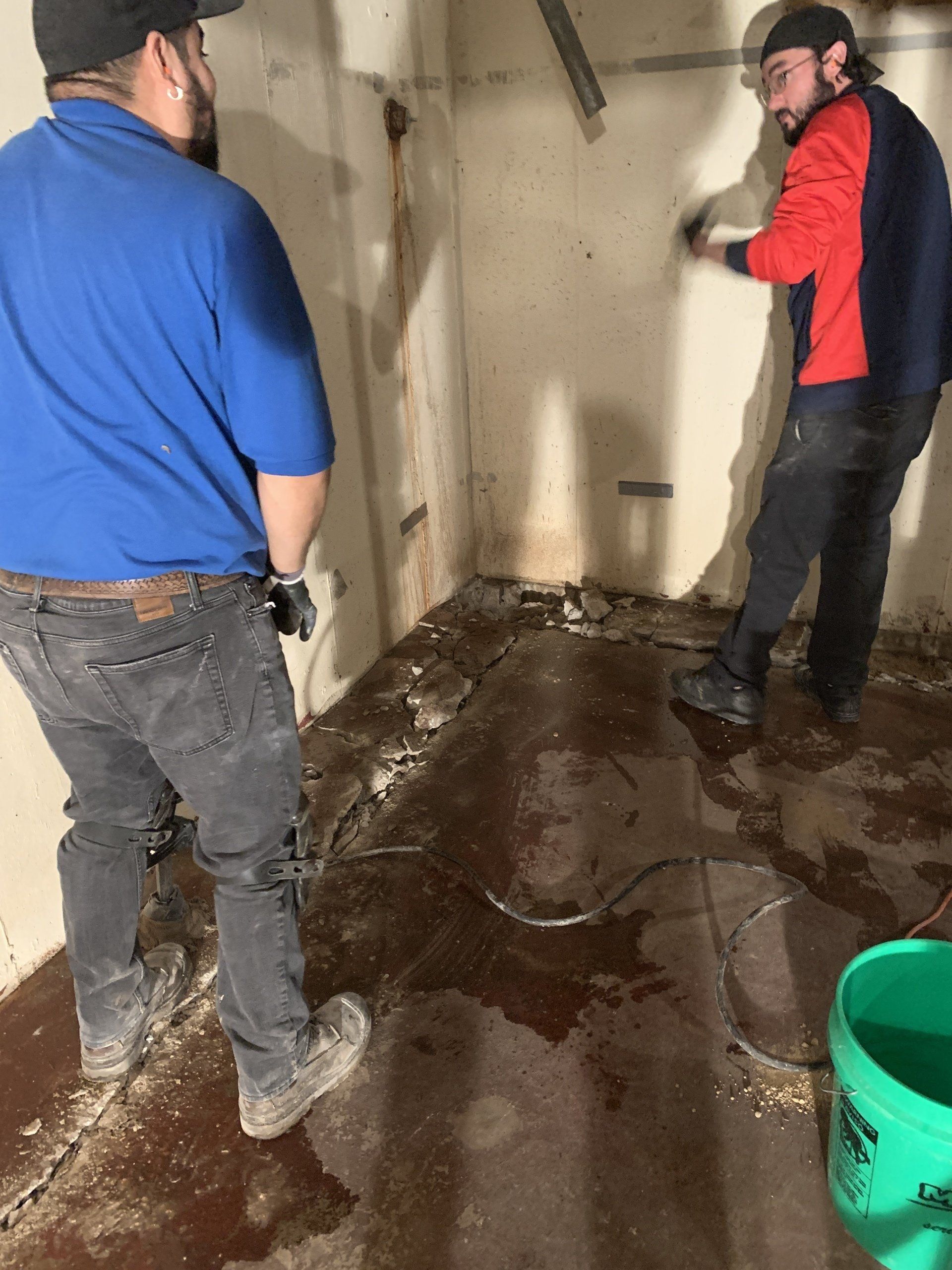Two men in a damp basement with a water-damaged floor. One points, and the other looks on near a bucket.