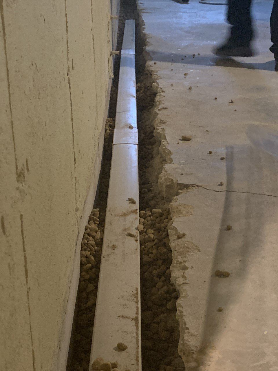 A narrow trench cut into a concrete floor next to a wall. A metal channel runs along the trench. Someone walks by in the background.