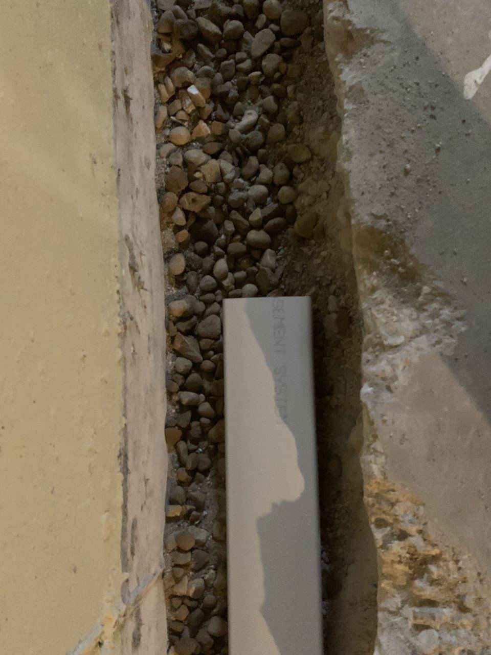 Gravel-filled trench with a white PVC pipe. The trench is between a tan wall and a grey concrete structure.
