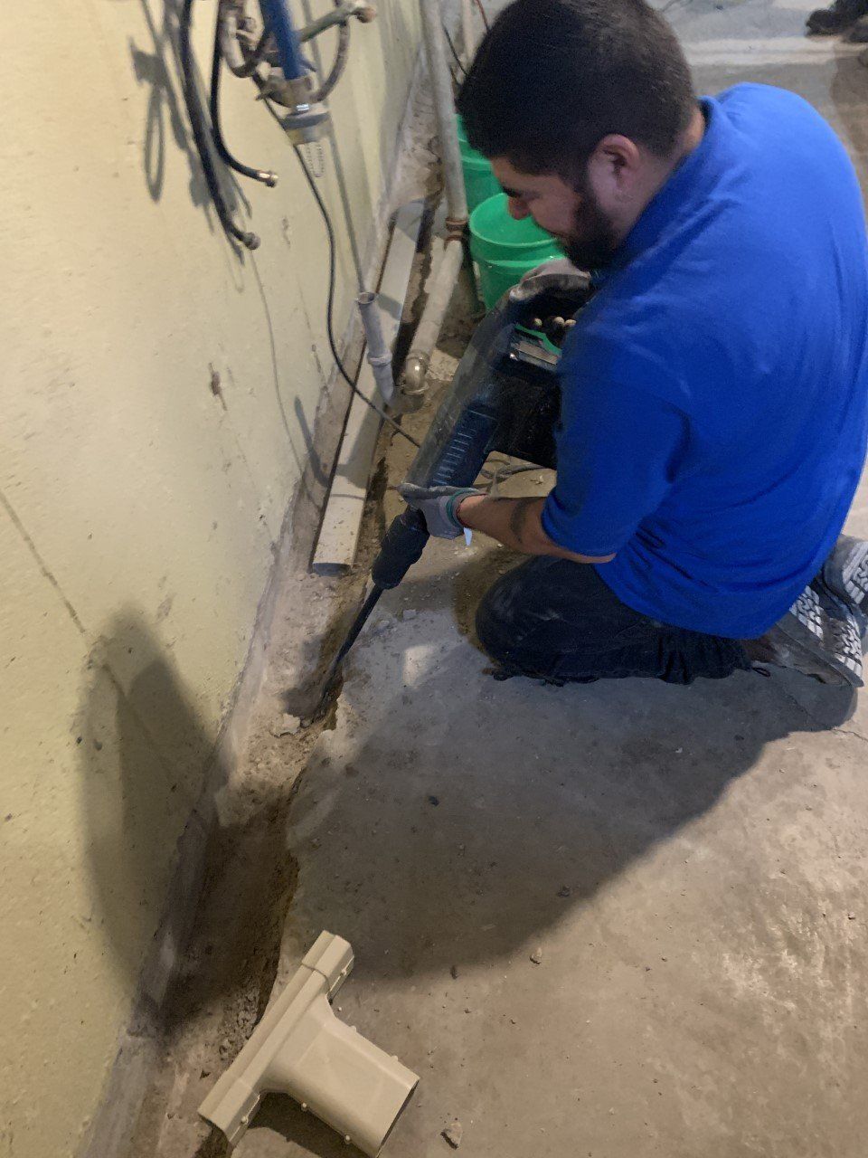 A man in a blue shirt kneels, working with a hose near a wall. The setting appears to be a basement.