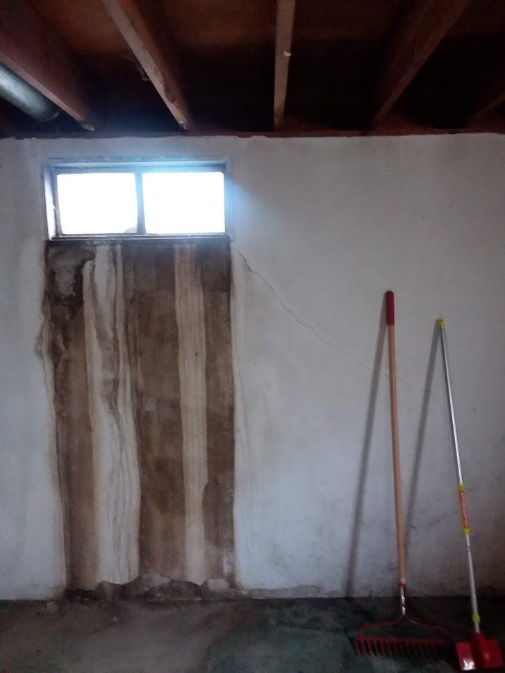 Basement wall with water damage around a small window. Two brooms lean against the wall.