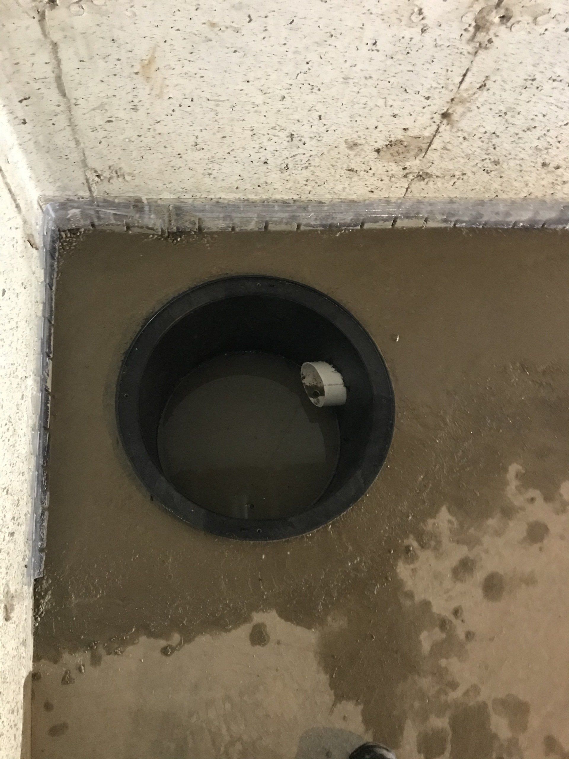 A dark, circular sump pit in a concrete floor filled with murky water. A white pipe enters the pit.