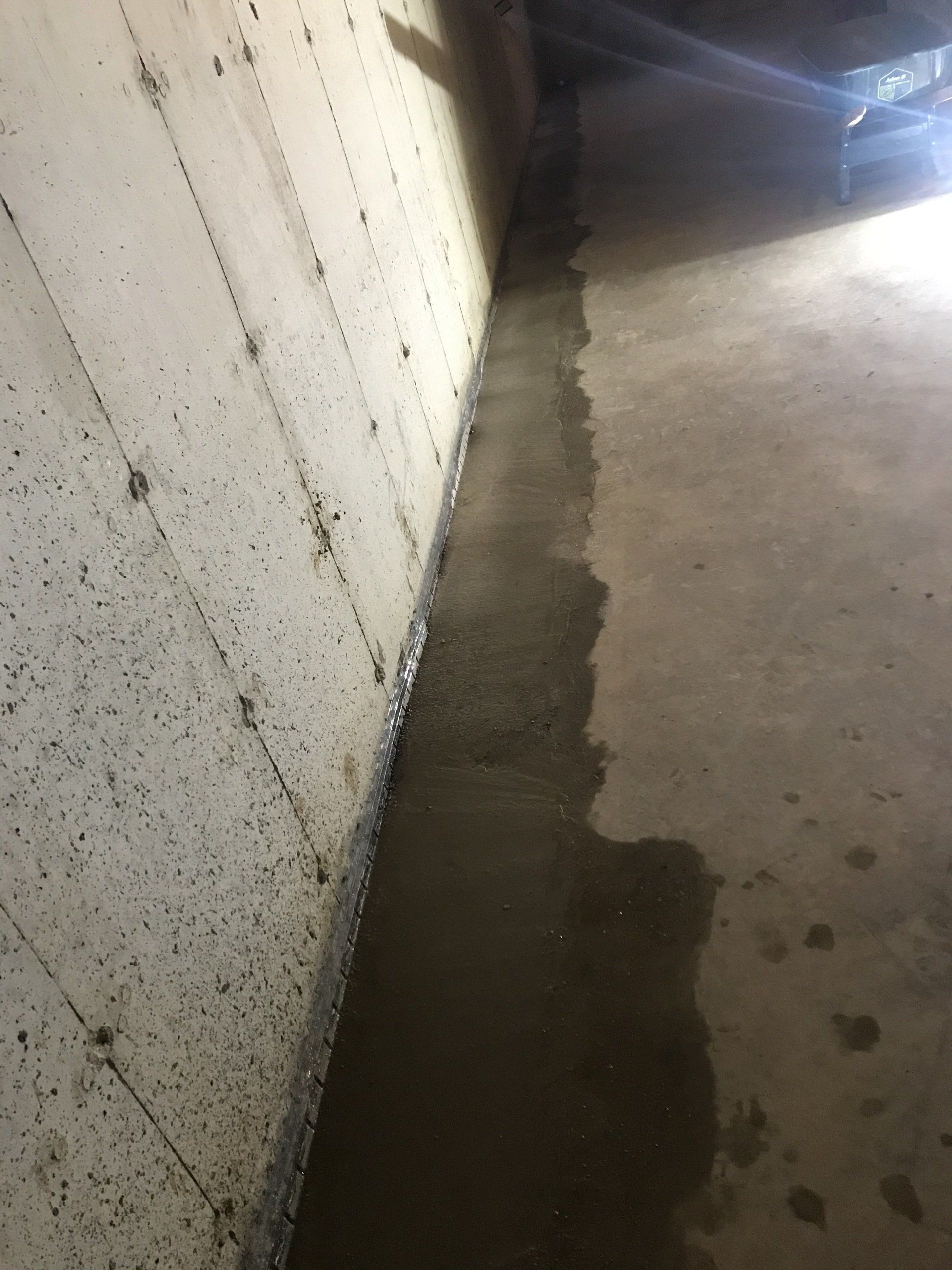 Water pooling along a concrete wall and floor in a basement. The concrete is gray and wet.