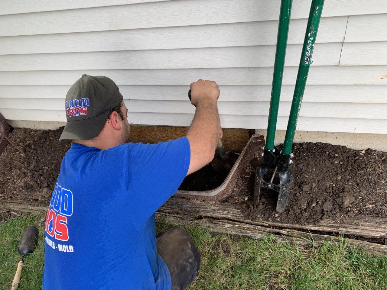 A man in a blue shirt digs near the foundation of a white house, using a shovel. Soil and a green handle tool sit nearby.