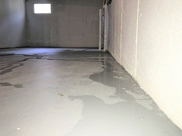 Flooded basement with standing water on a grey concrete floor, likely due to water damage. White walls and a small window are visible.