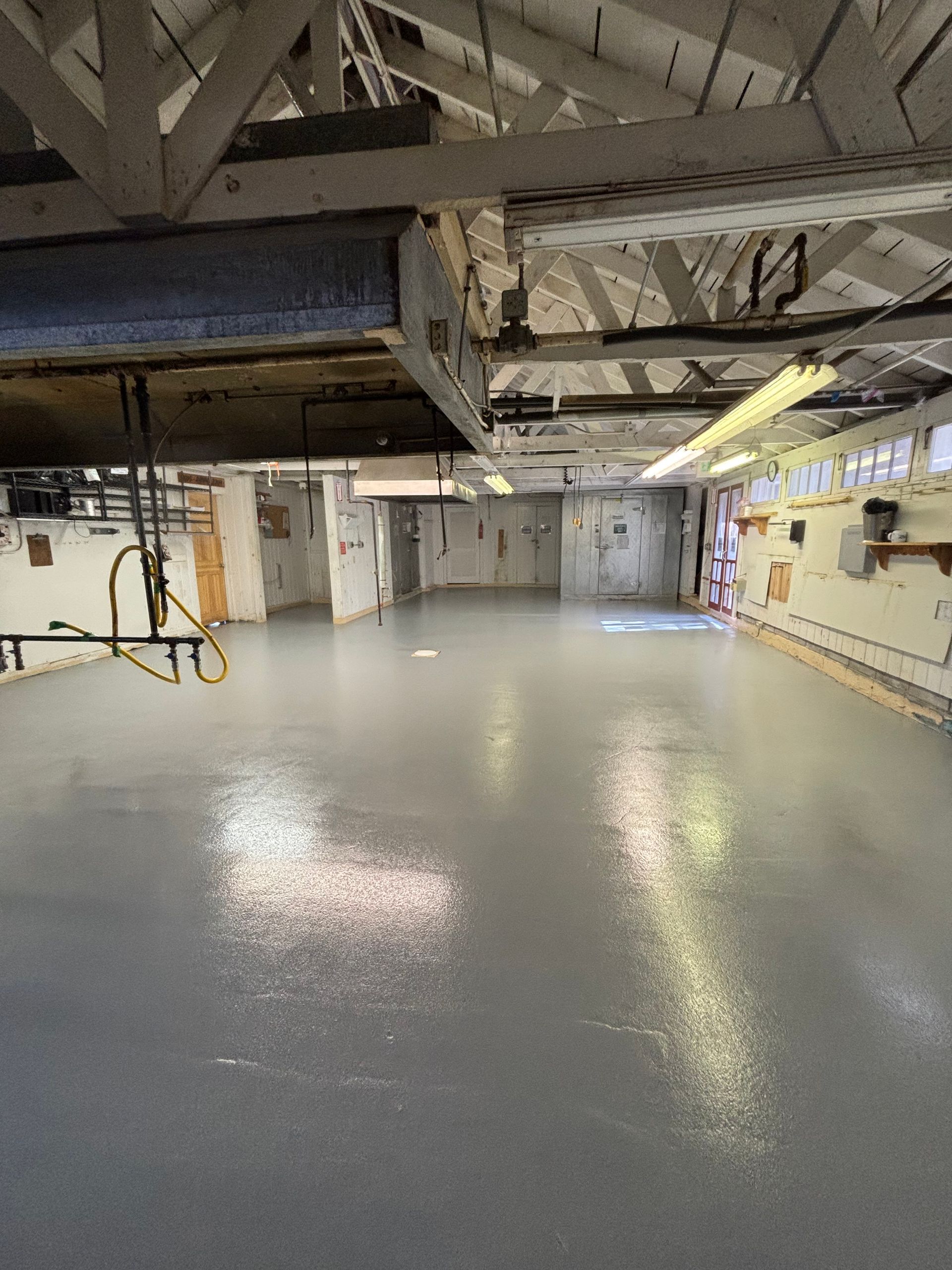 A spacious, freshly painted gray-floored basement with exposed rafters and overhead pipes.