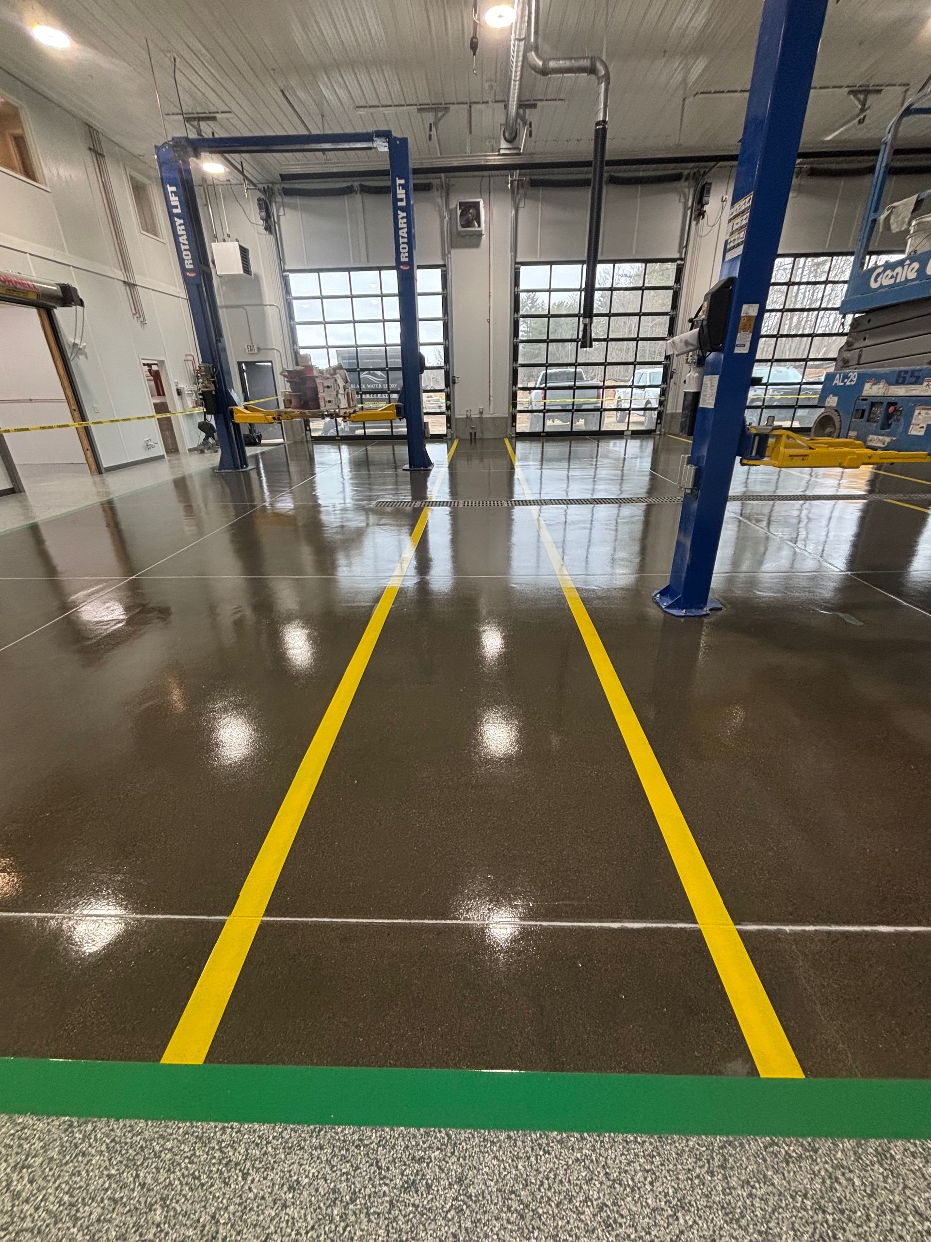 Yellow lines painted on a glossy gray floor in a garage. Two blue vehicle lifts, glass doors, and fluorescent lighting are visible.