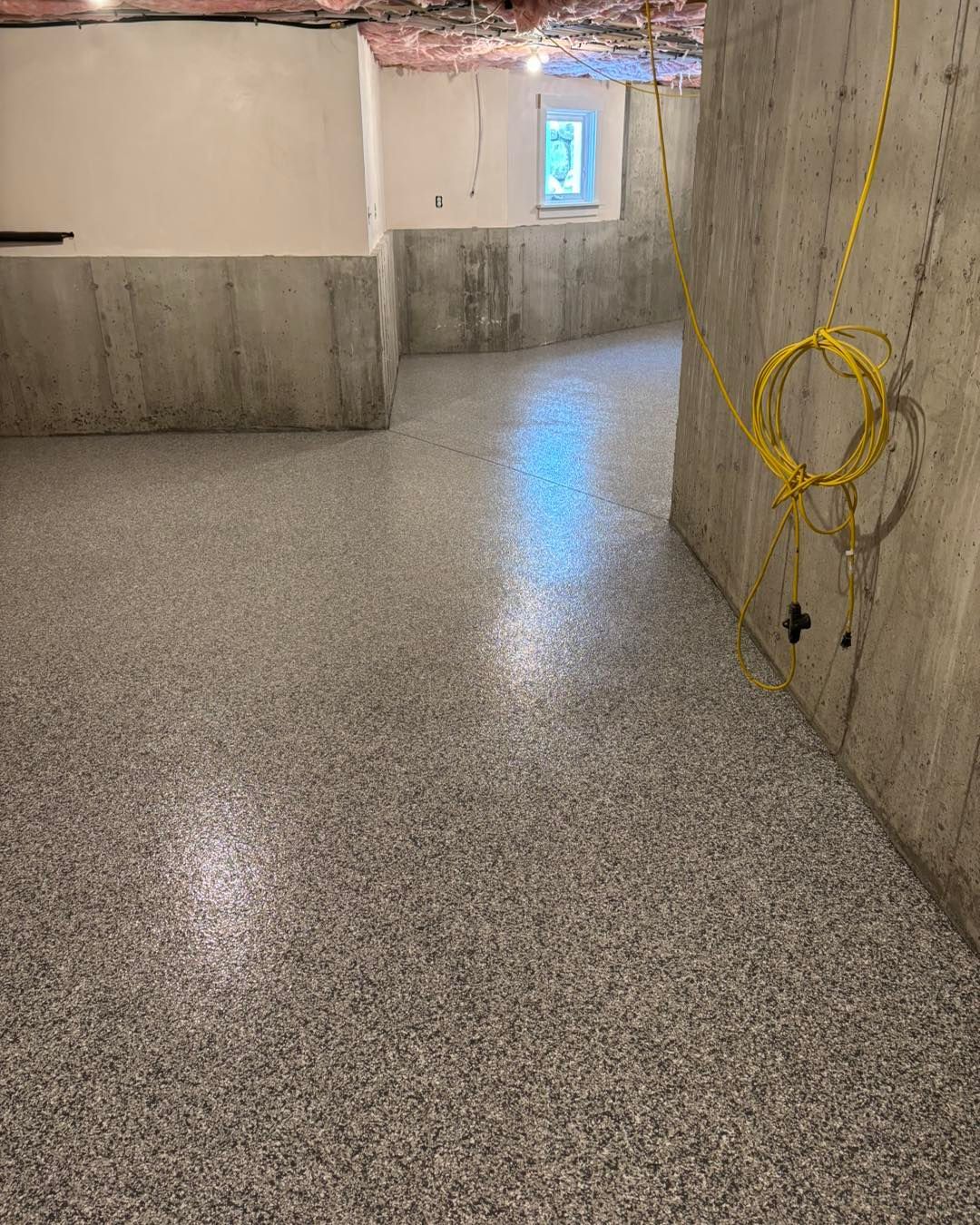 Basement with speckled gray flooring, concrete walls, and a window. A yellow electrical cord hangs on the wall.