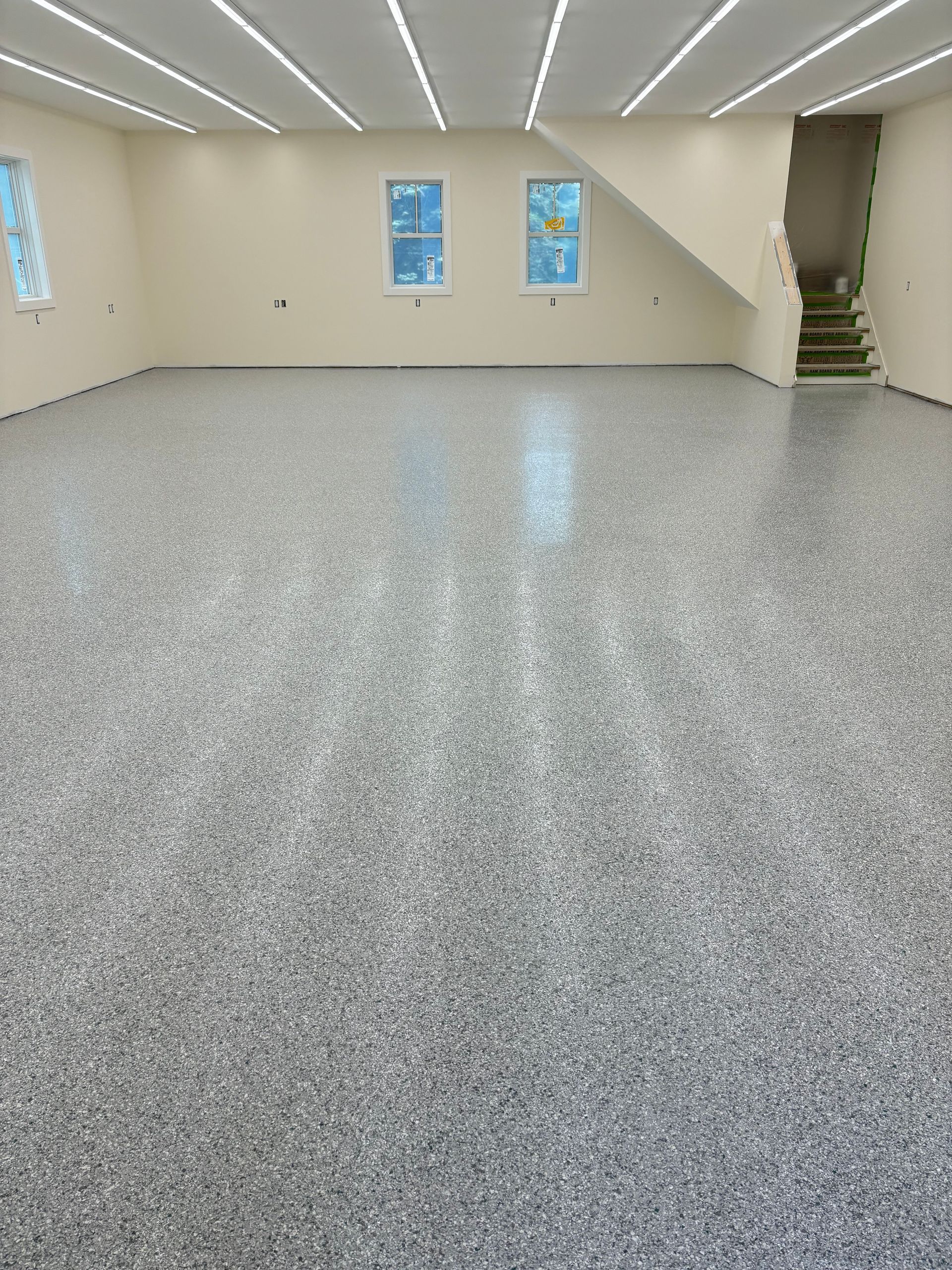 Empty room with epoxy floor, two windows, and staircase. Beige walls, fluorescent lighting.