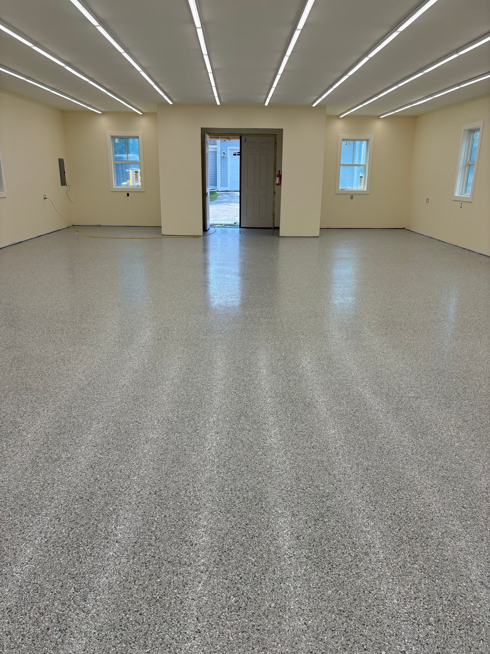 Empty room with speckled grey floor, beige walls, and bright overhead lights. Doorway leads outside.