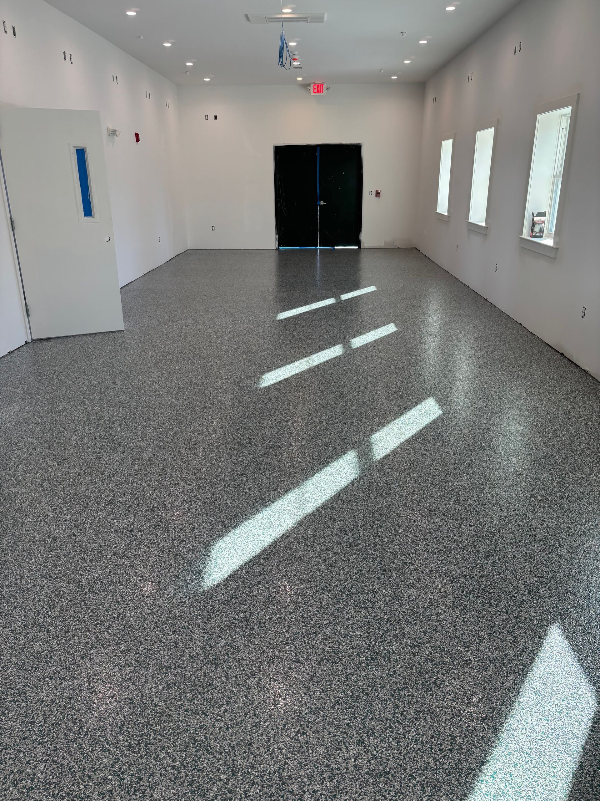 Empty room with speckled gray floor, white walls, black doors, and windows letting in sunlight.