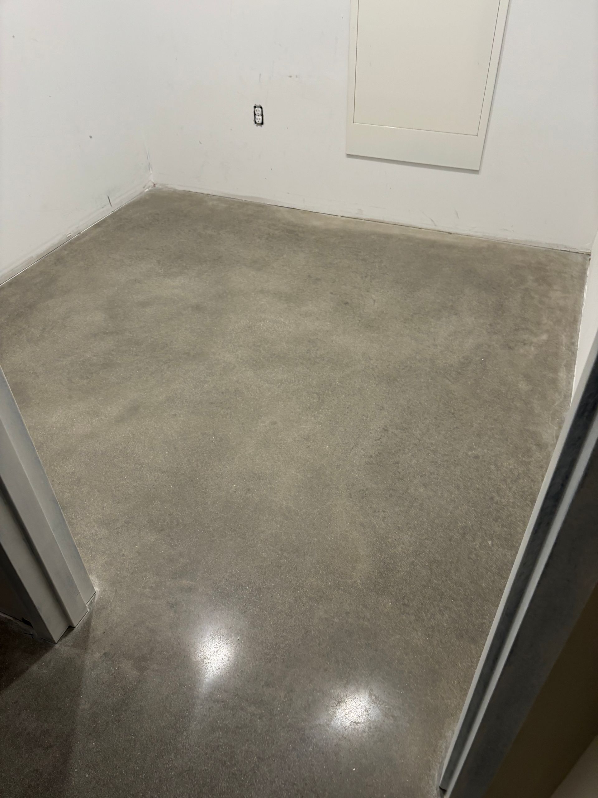 Gray concrete floor in a small, empty room with white walls and a small electrical outlet.