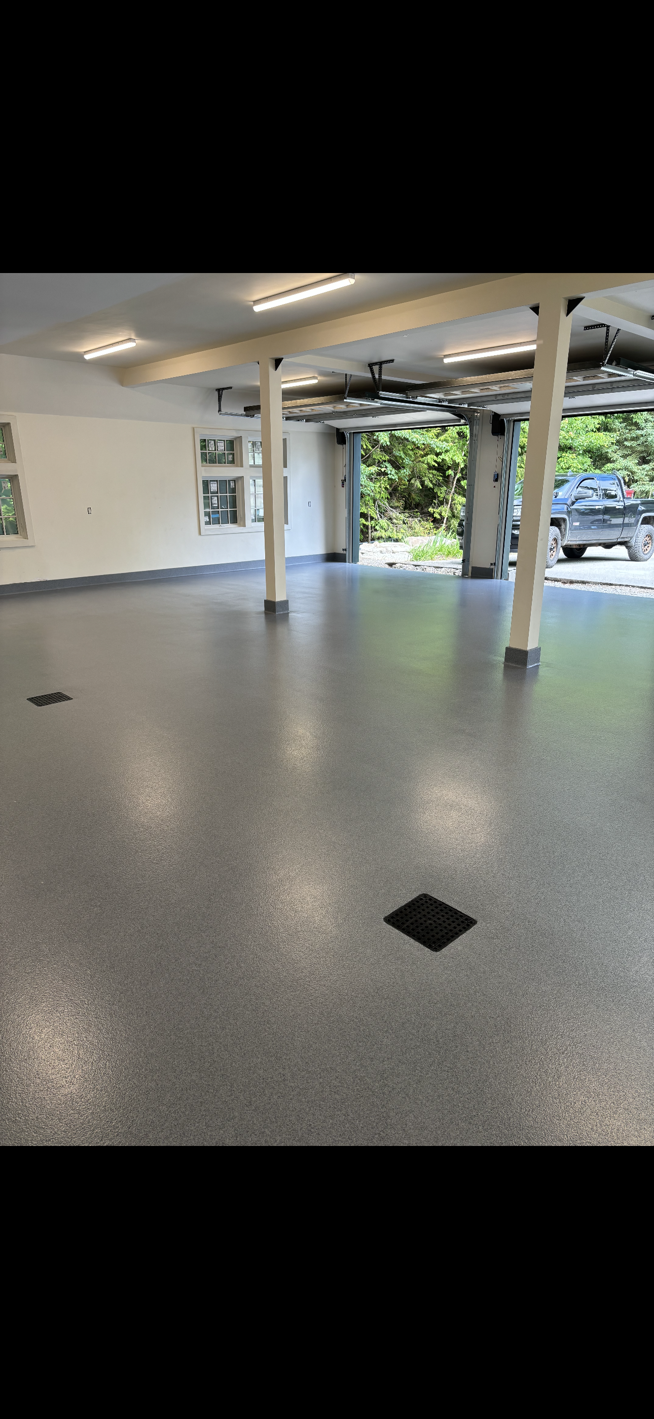 Empty garage with epoxy floor, open garage door leading to trees, white walls, and support beams.