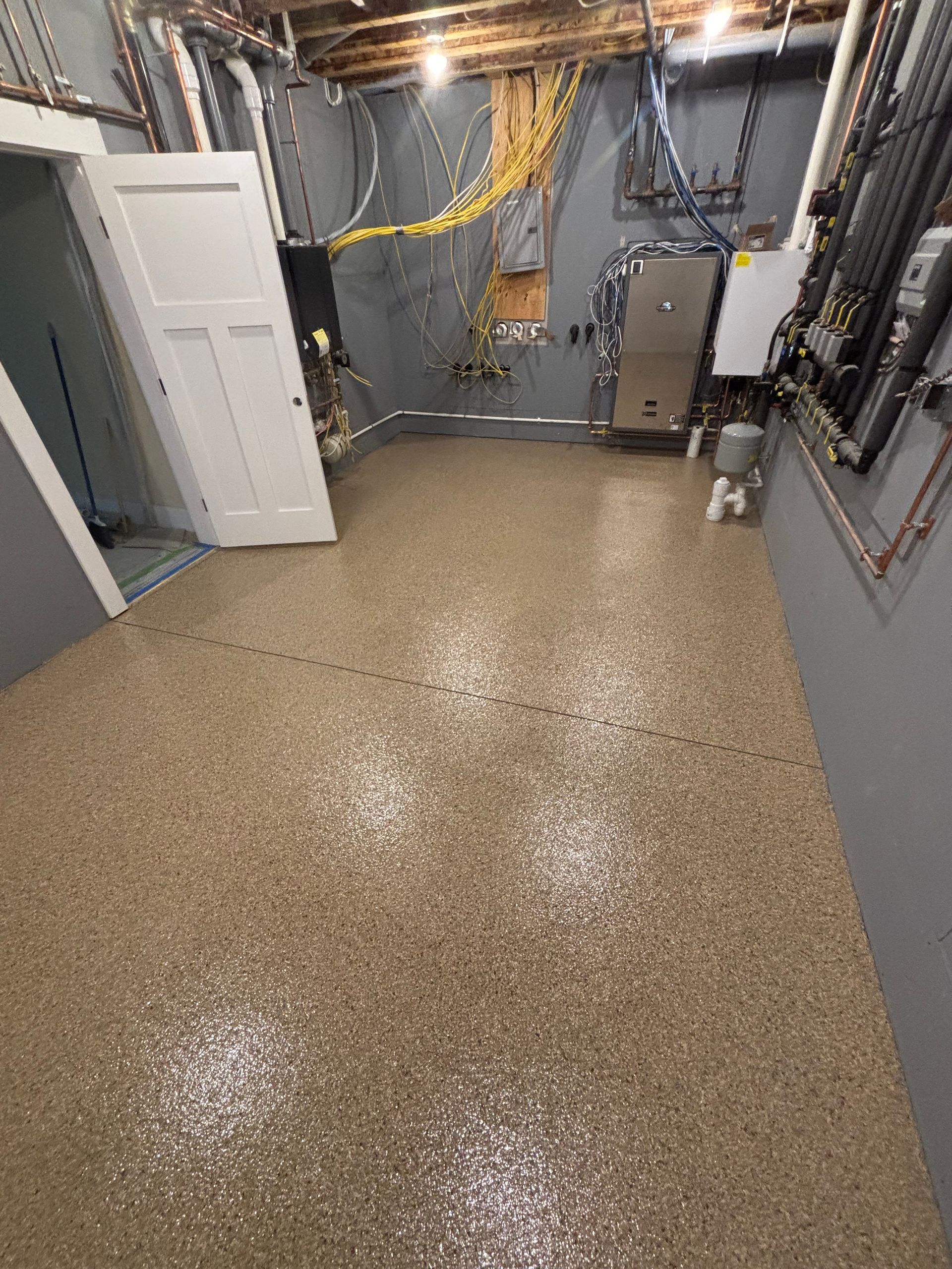 Basement with speckled flooring, grey walls, white door, and exposed pipes and wires on the walls.