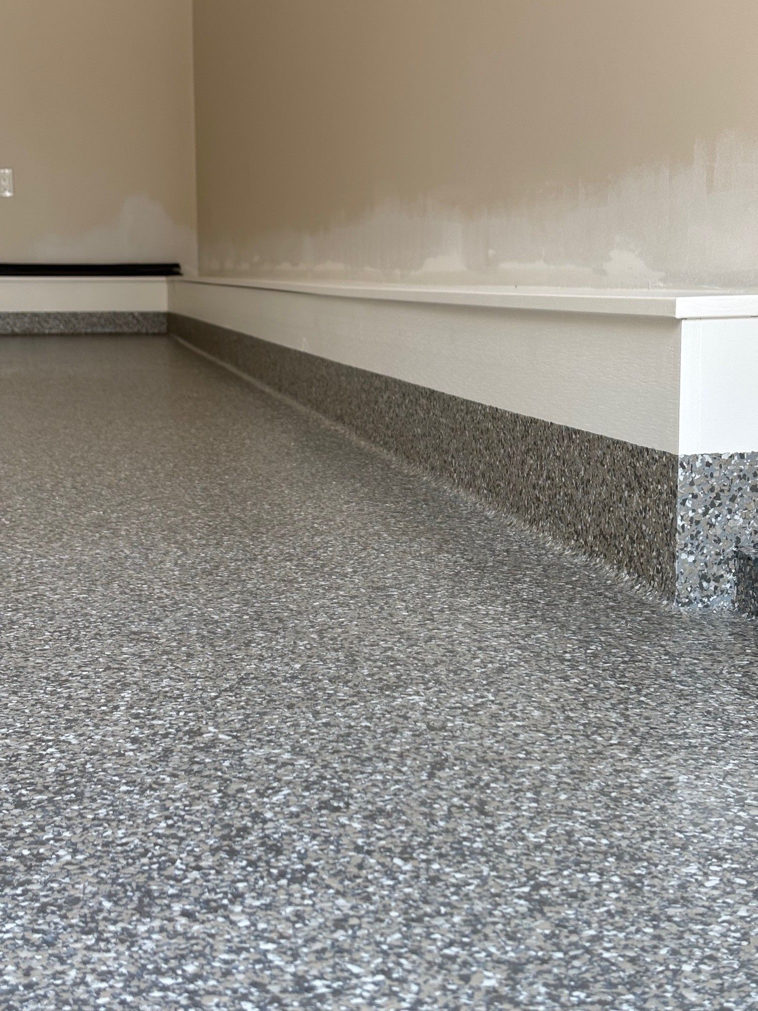 Epoxy-coated garage floor with gray flecks, bordering a white baseboard and tan wall.