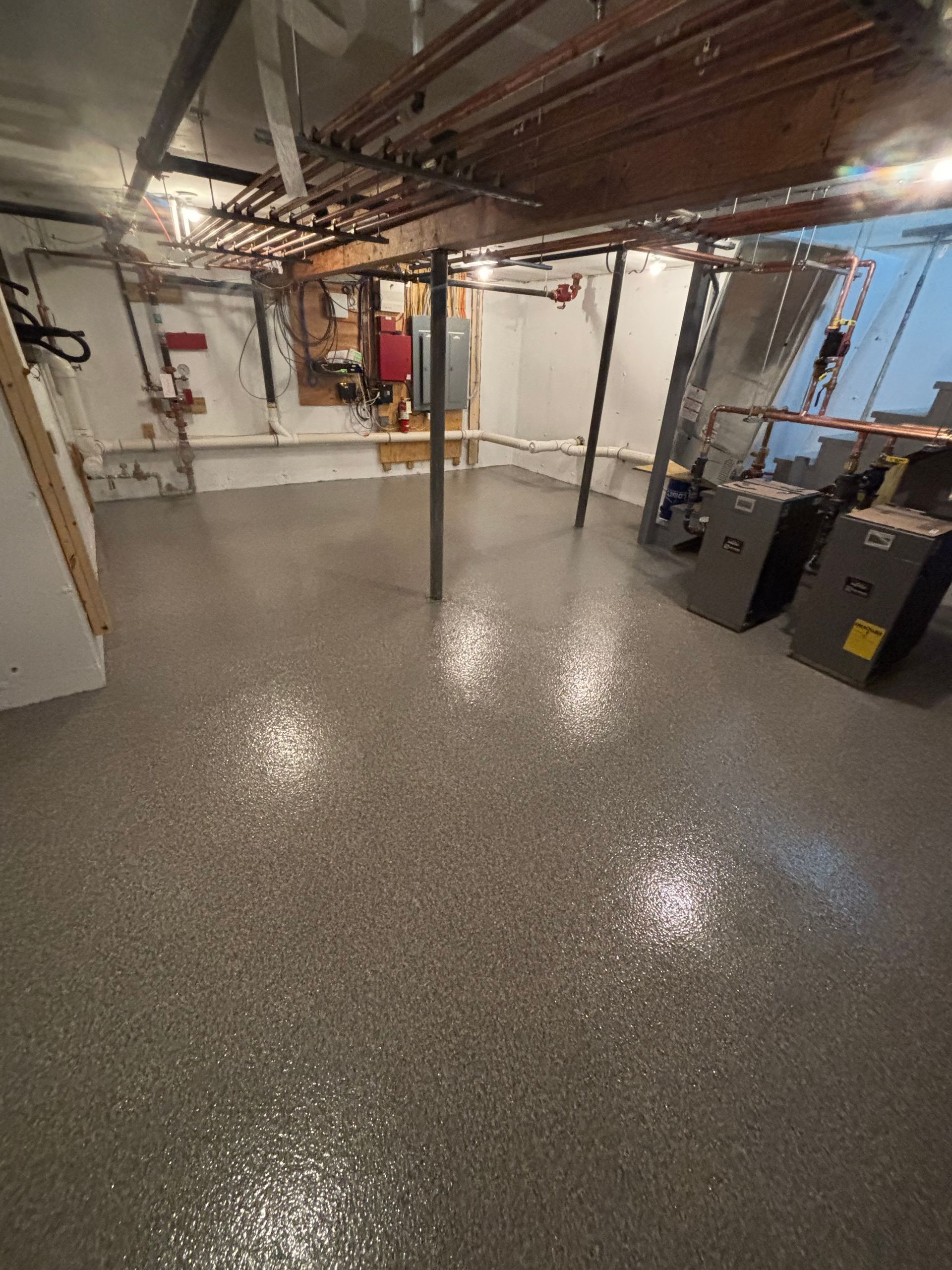 A basement with a shiny, speckled gray floor. Exposed pipes and support beams run along the ceiling.