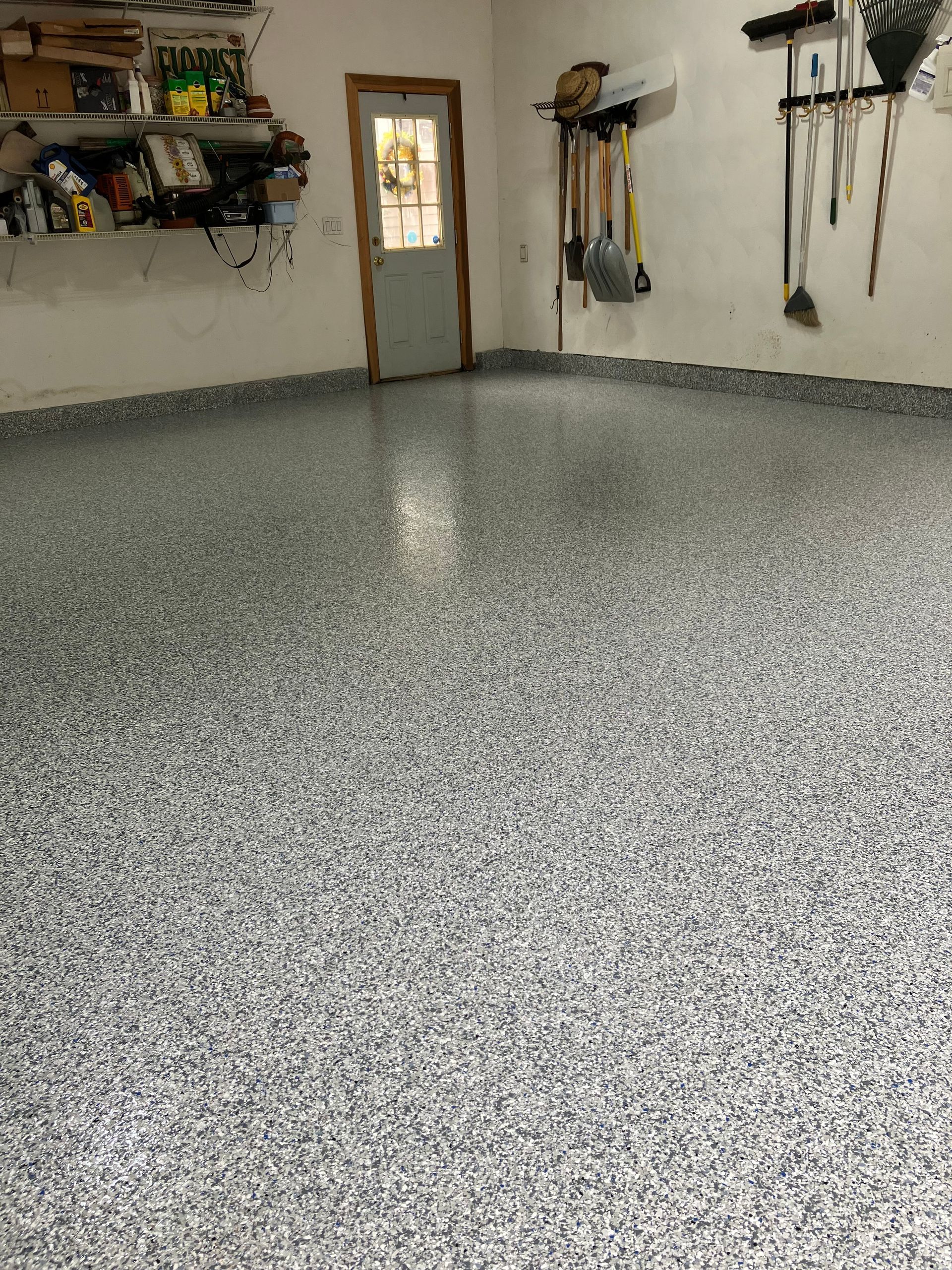 Garage with speckled gray epoxy flooring, shelves with items, door, and tools hanging on the wall.