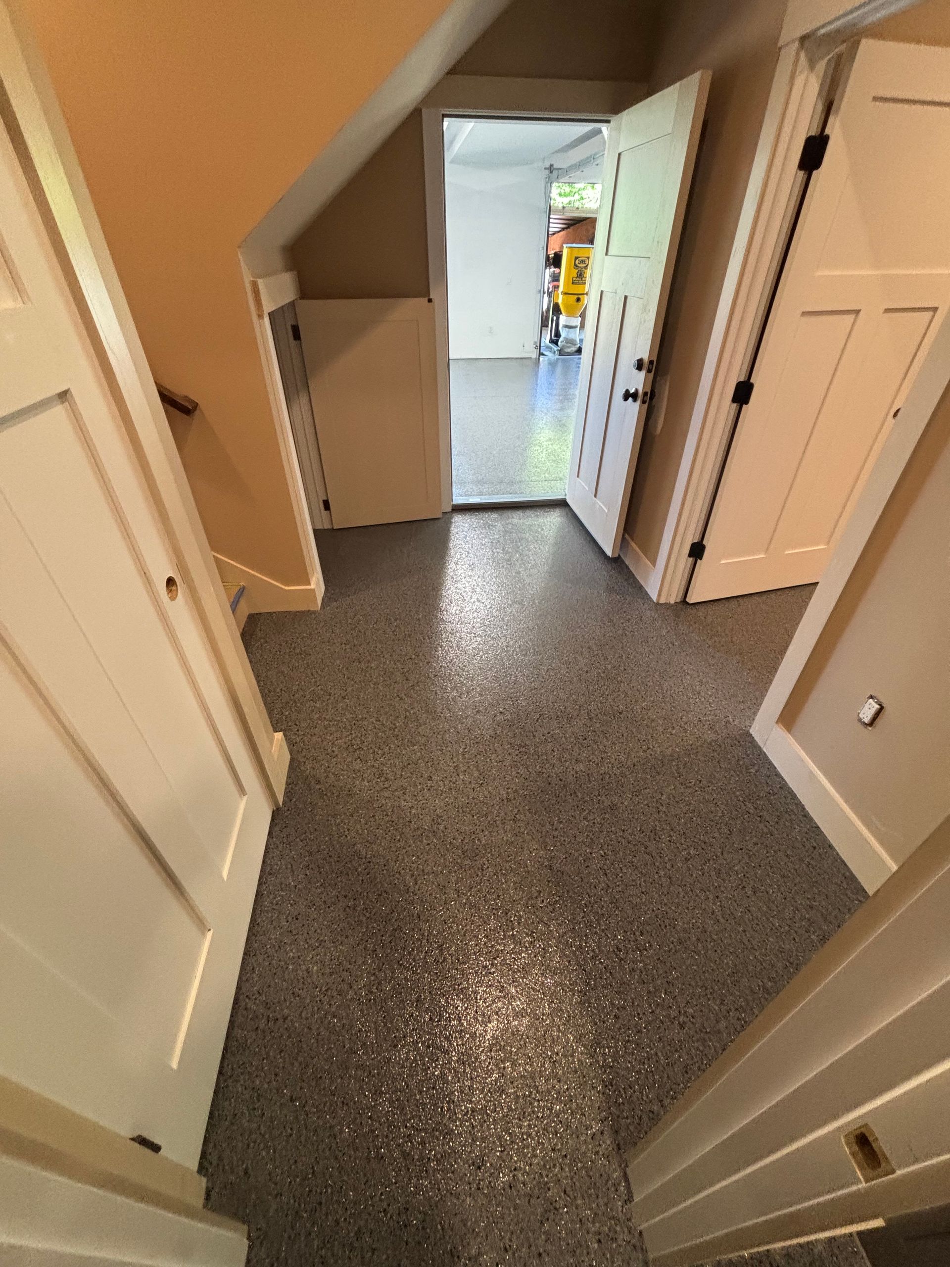 Hallway with speckled gray flooring, white doors, and a doorway leading to a brightly lit room.