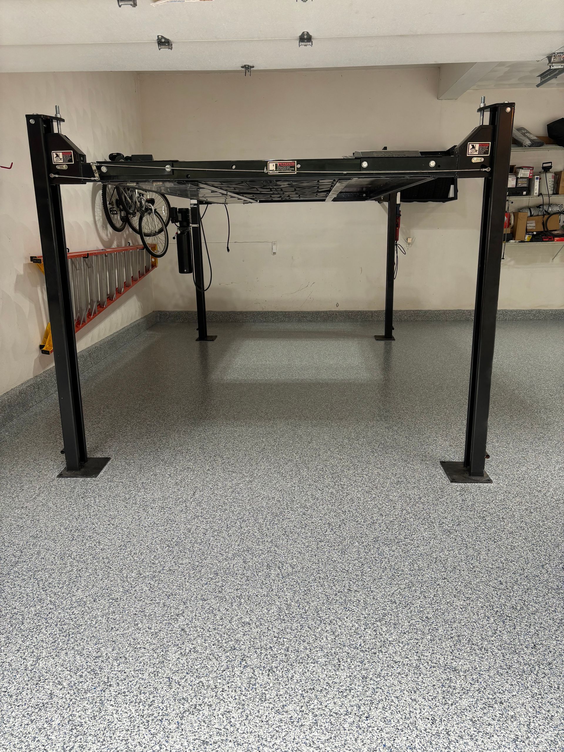 Four-post car lift in a garage with a speckled floor. The lift is black with no vehicle on it.