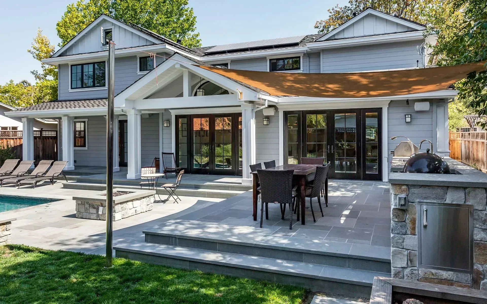 A light-gray two-story house features a stone patio with outdoor dining, a fire pit, lounge chairs, and a pool.